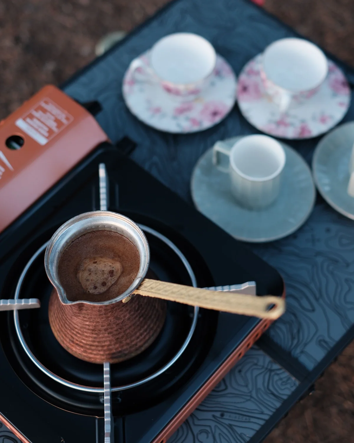A copper coffee pot on a portable gas stove, with cups ready beside it